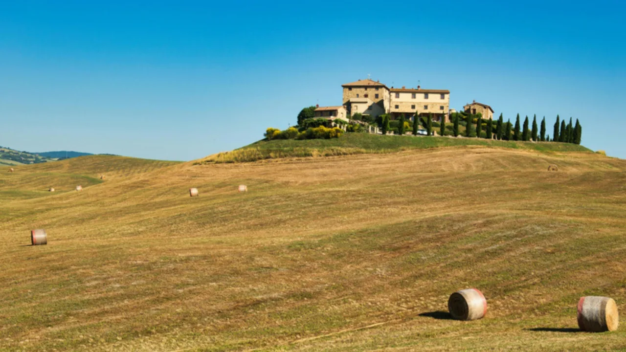 A stone farmhouse and cypress trees on a hill above harvested fields in the Tuscan countryside